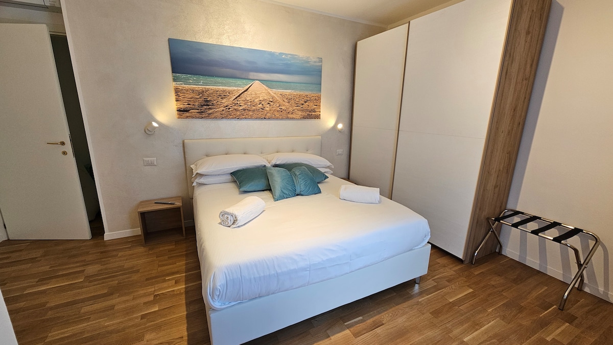 A well-lit bedroom features a spacious bed with decorative pillows and fresh towels arranged neatly. A large piece of beach-themed artwork enhances the room’s ambiance. A wardrobe and a folding stool are positioned to maximize space, complemented by warm wood flooring.