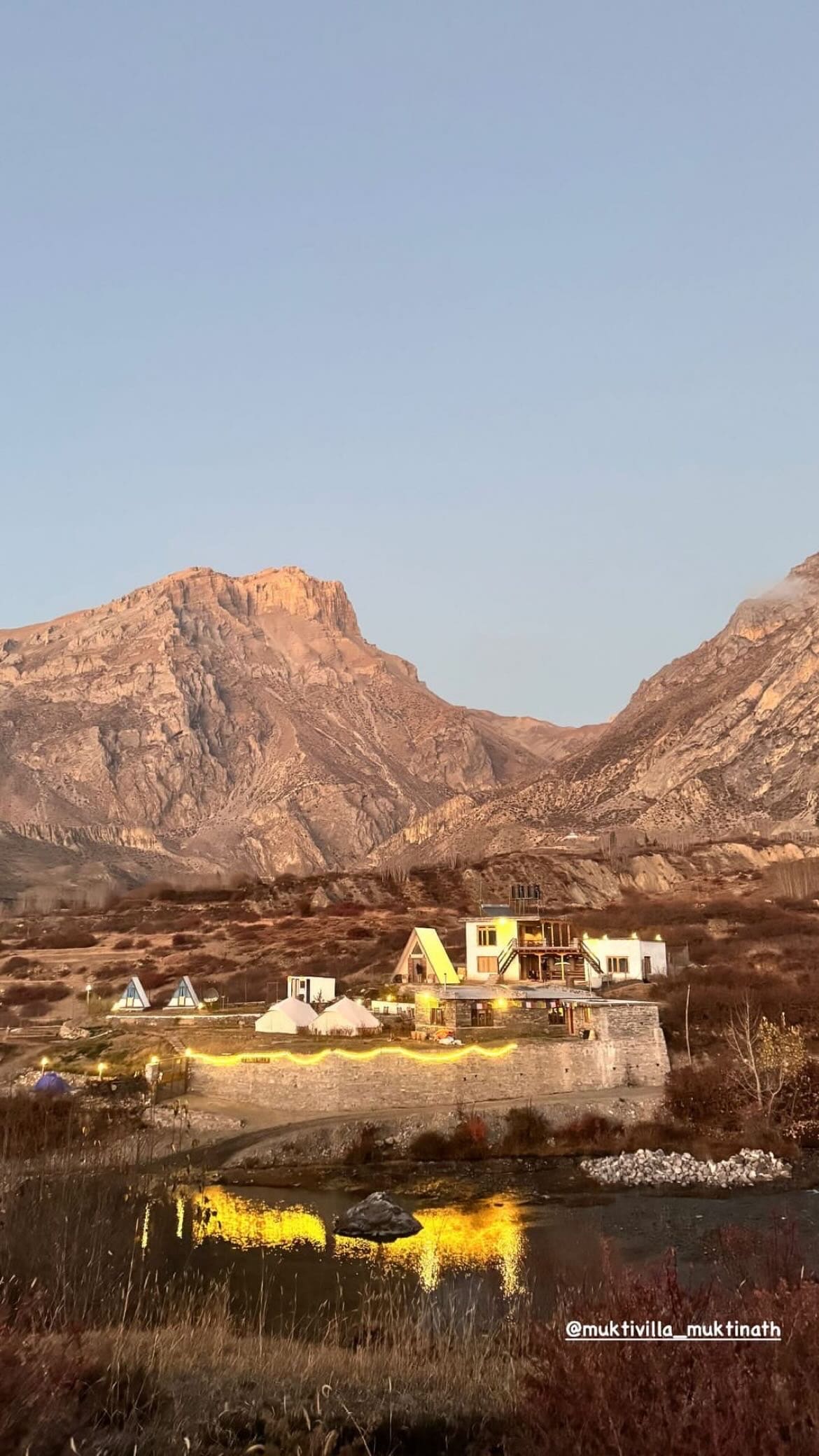 A charming building is nestled among rugged mountains, reflecting warm light from its exterior. The landscape includes hints of natural vegetation and a water body that complements the serene environment, while the mountains rise majestically in the background.