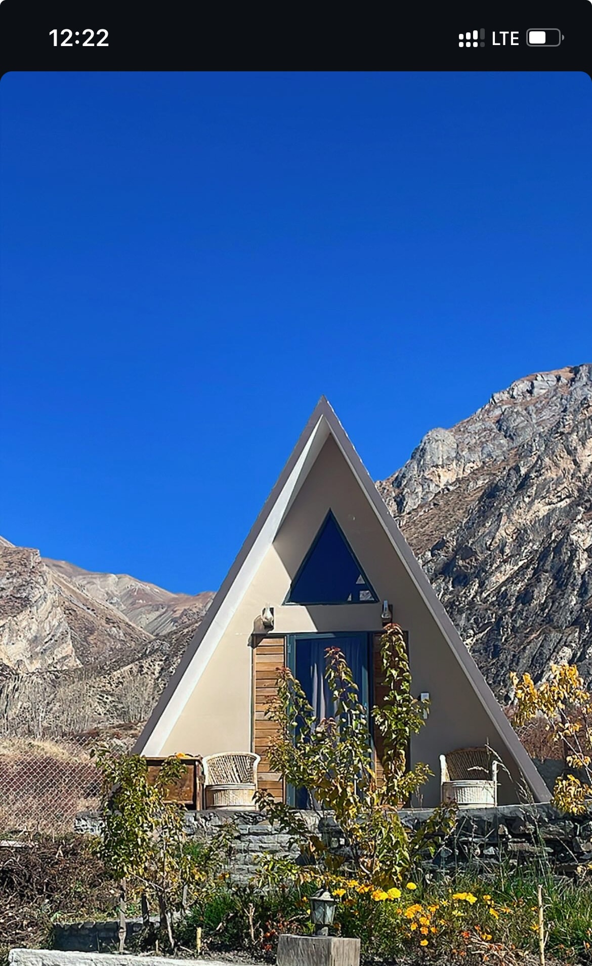 An A-frame structure is set against a clear blue sky, surrounded by rugged mountains. The entrance is adorned with a wooden door and two wicker chairs positioned on a stone platform, complemented by seasonal plants in the foreground.