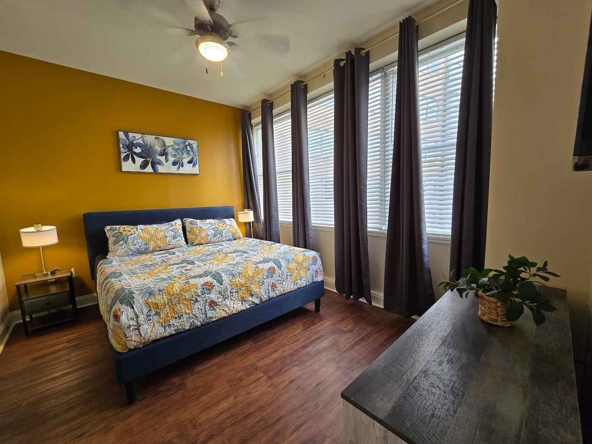 A bright bedroom features a spacious bed with a floral quilt, complemented by two bedside lamps on metal tables. Large windows with light-filtering shades allow natural light to fill the room. Dark curtains provide privacy, and a small plant is displayed on a wooden dresser.