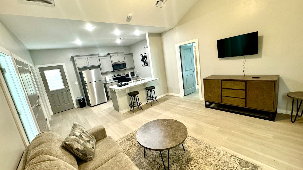 A spacious living area features light-colored wood flooring and high ceilings. A comfortable sofa is positioned near a circular coffee table, while two bar stools are placed at a kitchen island in the background. A mounted flat-screen TV is visible on a wall opposite a modern media console.
