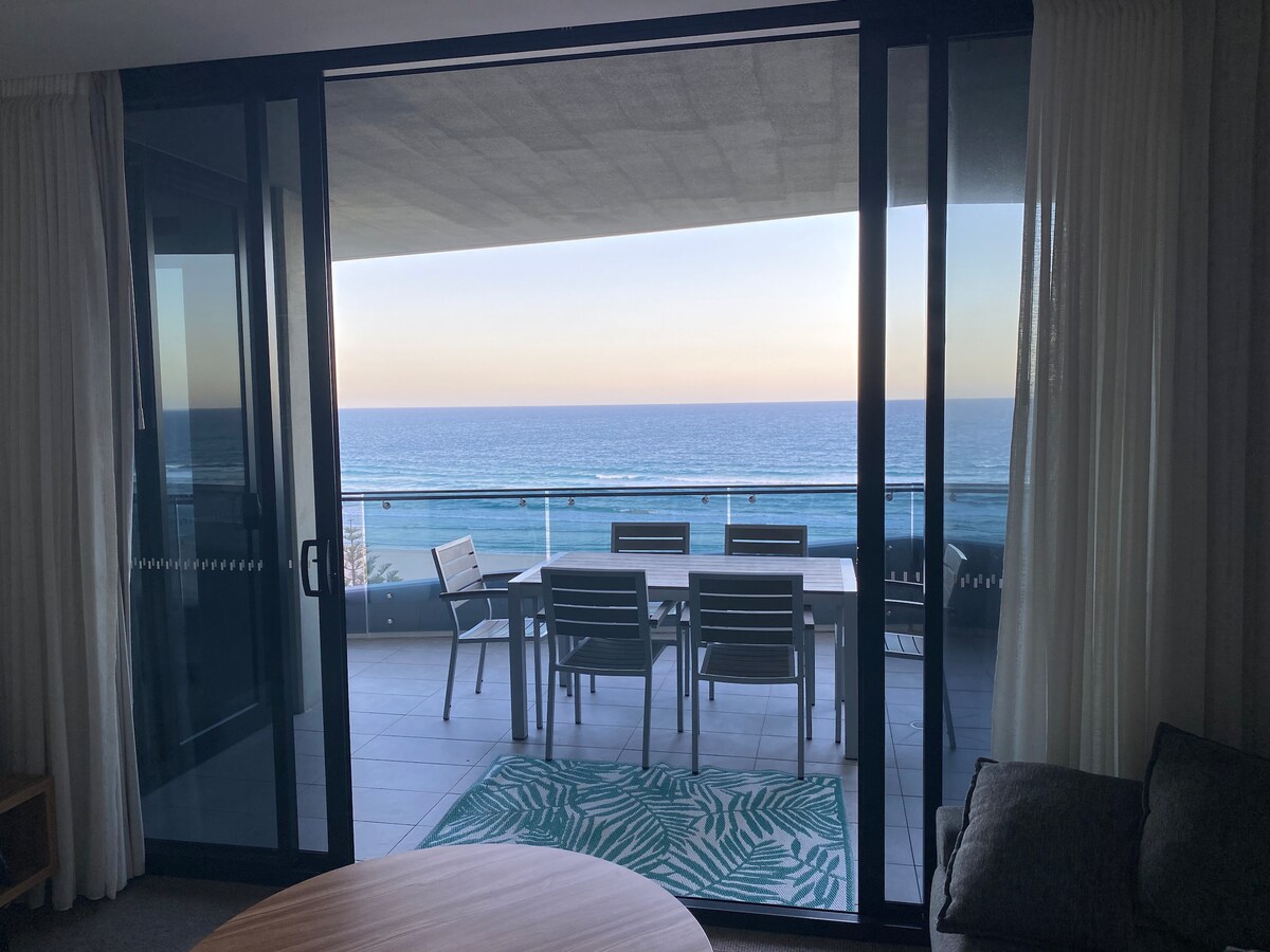 A spacious balcony is visible through floor-to-ceiling glass doors, showcasing a dining table and chairs set against the backdrop of the ocean. Soft, light curtains frame the view, while a patterned rug adds a touch of color to the outdoor space.