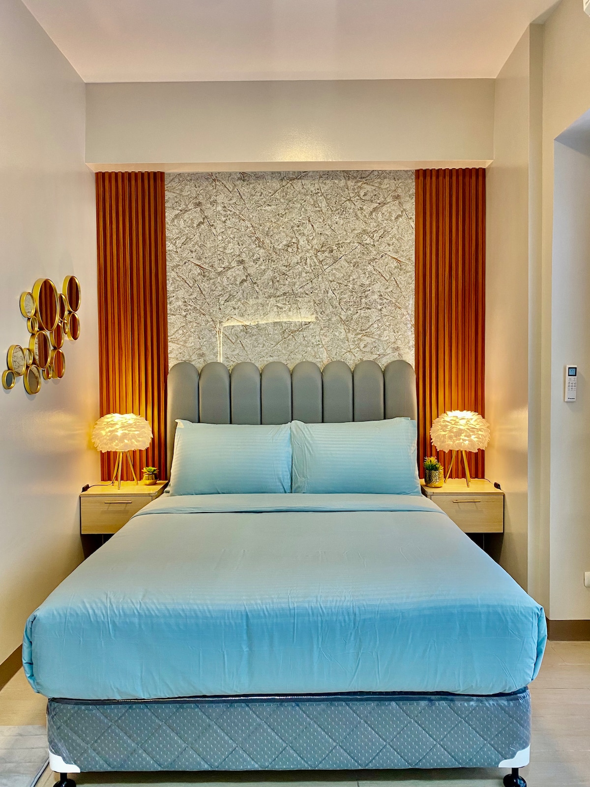A cozy sleeping area is showcased with a spacious bed covered in a soft blue blanket. On either side of the bed, modern bedside tables with unique lamps provide ambient lighting. Textured wall accents create a contemporary feel in the bright room.