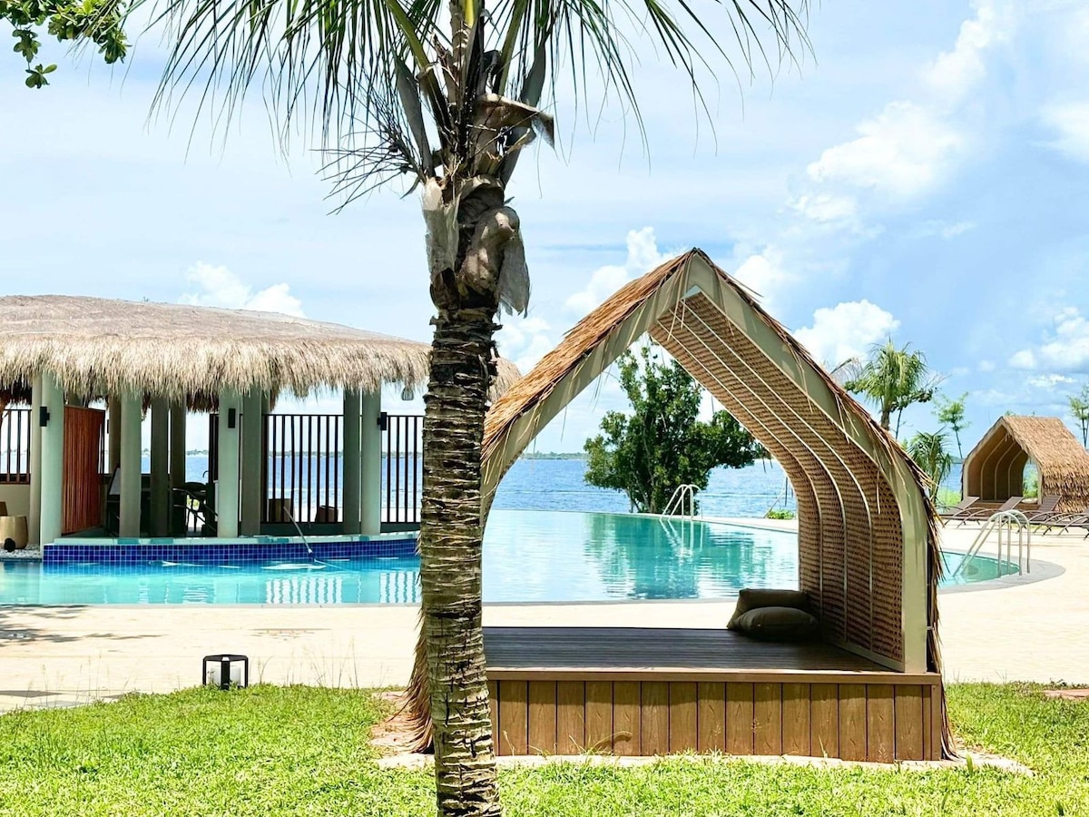 A shaded seating area is positioned beside a large pool, with views of the water in the distance. Lush greenery surrounds the space, and a palm tree provides natural shade. The pool features a gentle curve and is accompanied by a thatched-roof cabana.