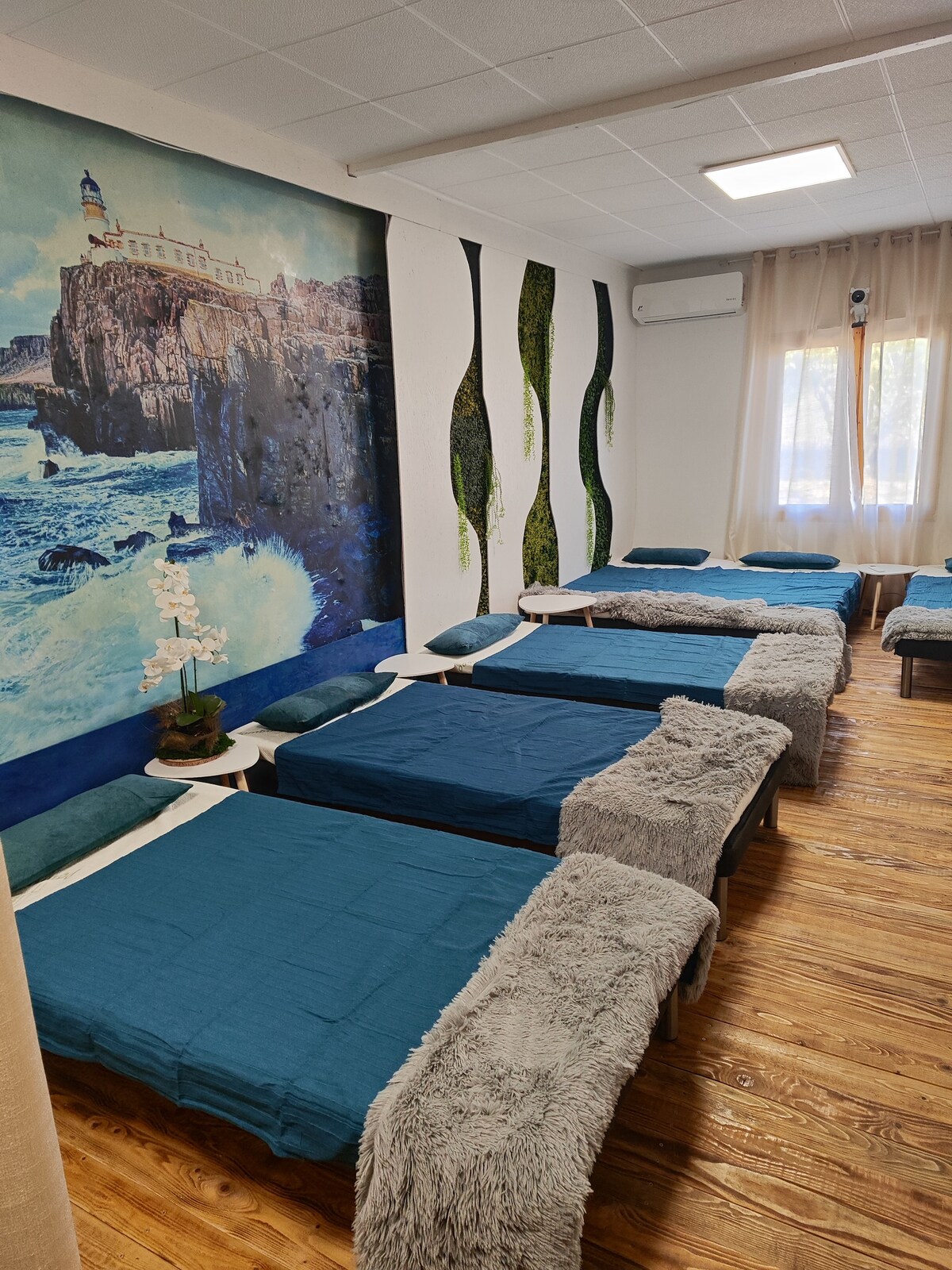 A spacious sleeping area is displayed with several neatly made beds, each covered in blue linens. A mural of a coastal scene adds a serene touch to the wall. Natural light filters through the window, enhancing the calming atmosphere of the room.