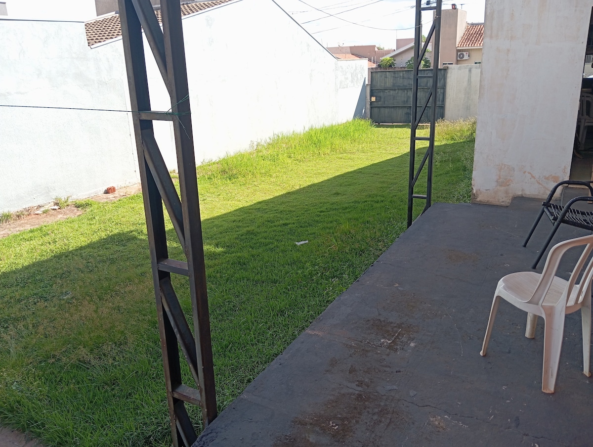 A spacious outdoor area is seen, featuring a green lawn bordered by concrete walls. A covered patio with a solid floor indicates a suitable space for relaxation. Two chairs are positioned near the patio edge, inviting leisurely moments in the tranquil setting.