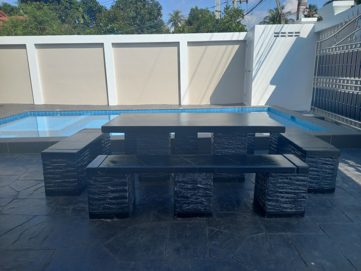 A dining area features a large black stone table surrounded by matching benches, set beside a swimming pool. The pool is visible in the background, with clear blue water contrasting the neutral tones of the surrounding walls.