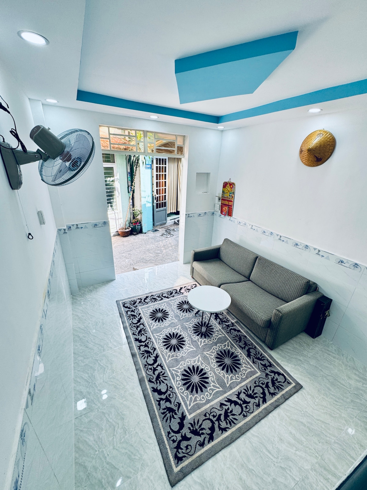 A modern living area features a comfortable gray sofa positioned beside a small round table. A decorative rug is placed on the shiny tiled floor, while natural light enters through an open doorway leading to an outdoor space.