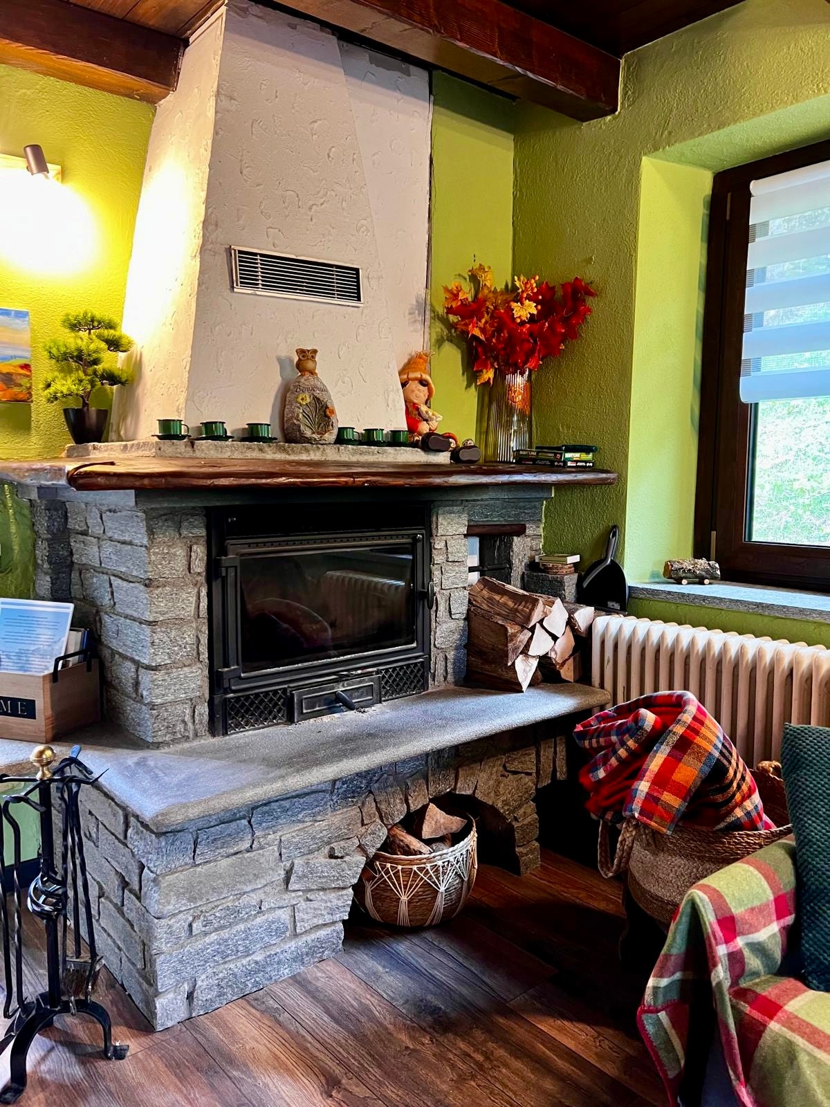 A stone fireplace features a modern insert and a rustic wooden mantle adorned with decorative items. Nearby, a comfortable seating area is complemented by a cozy blanket, adding visual warmth to the space. Soft light filters through a window, enhancing the inviting atmosphere.