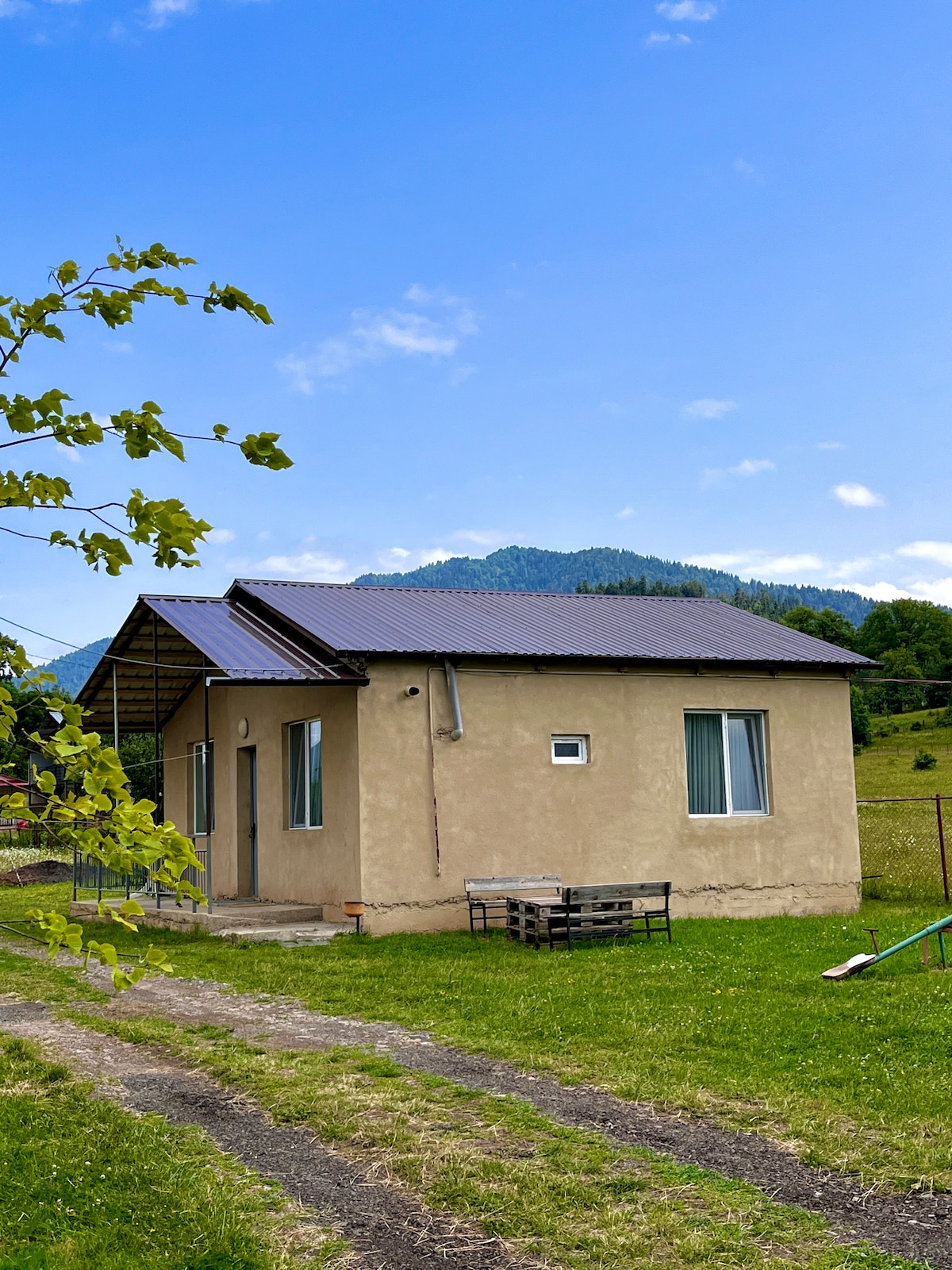 A cottage is set against a backdrop of rolling green hills and a clear blue sky. The single-story structure features a sloped roof, two large windows, and a small porch area. Grass surrounds the home, providing a natural setting.