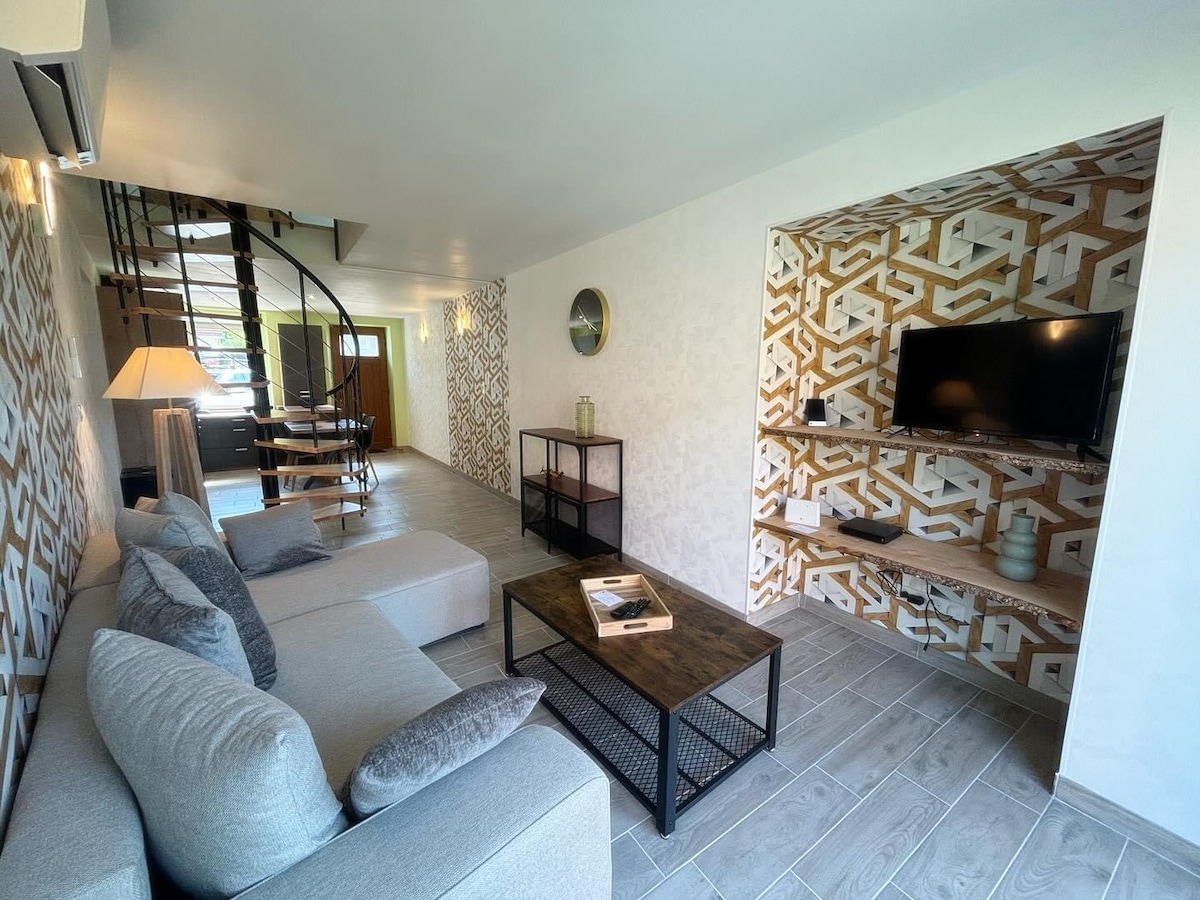A modern living area is presented with a light-colored sectional sofa adorned with decorative pillows. A dark coffee table with a metal frame sits in the center. The wall features a geometric design, while a flat-screen television is mounted on a nearby display unit. Natural light enters through a spiral staircase leading to an upper level.