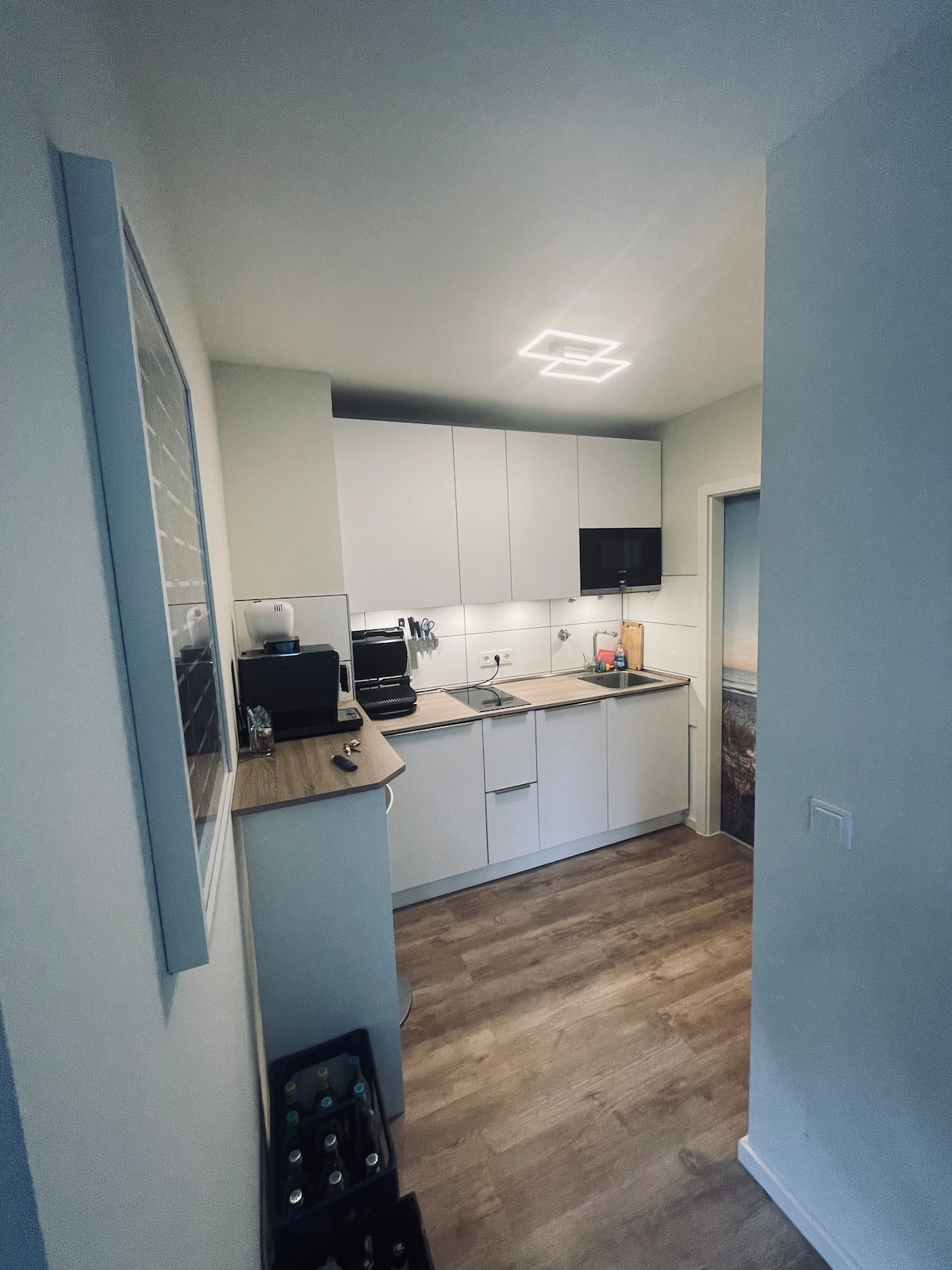 A modern kitchen is equipped with sleek white cabinets and essential appliances, including a coffee maker and microwave. A small window allows natural light to brighten the space, while a lighted ceiling fixture adds illumination. The flooring features a wood-like design, enhancing a clean aesthetic.