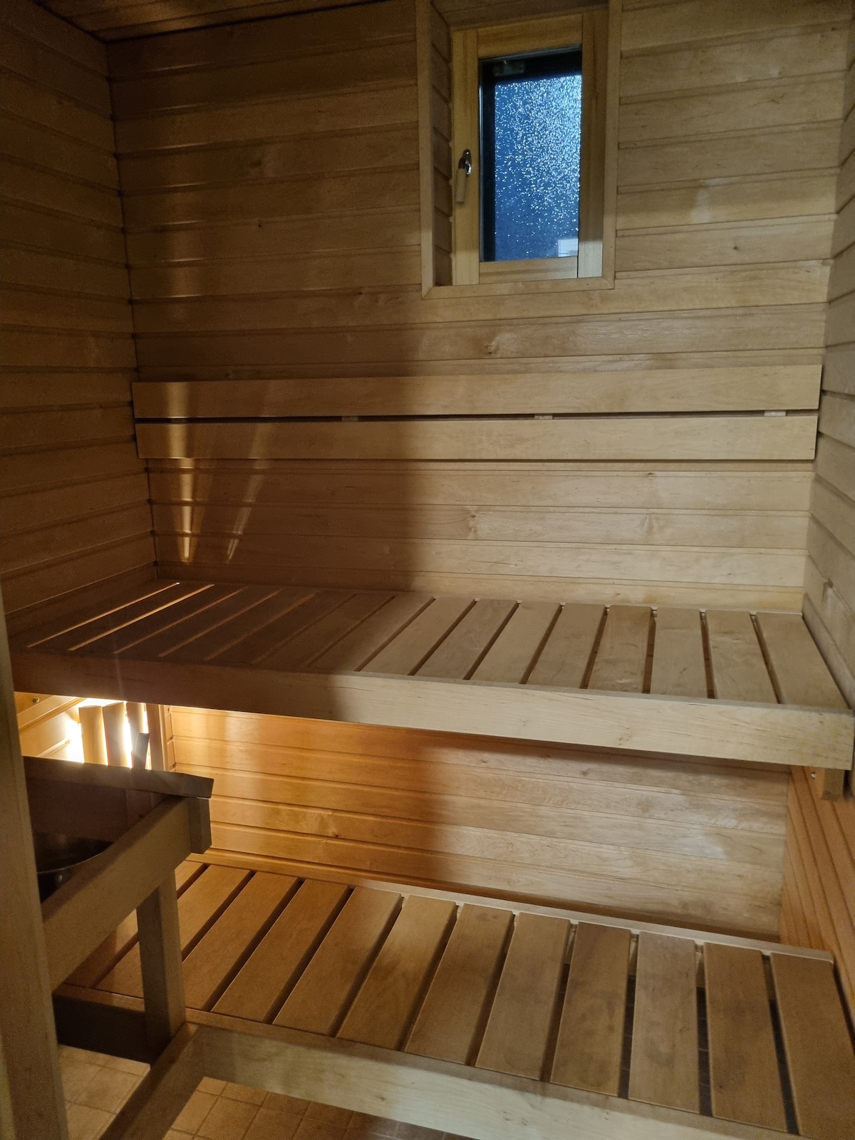 A wooden sauna interior is presented with two tiered benches. Natural wood paneling lines the walls, creating a warm and inviting space. Light filters in through a small window, adding a subtle glow to the atmosphere.