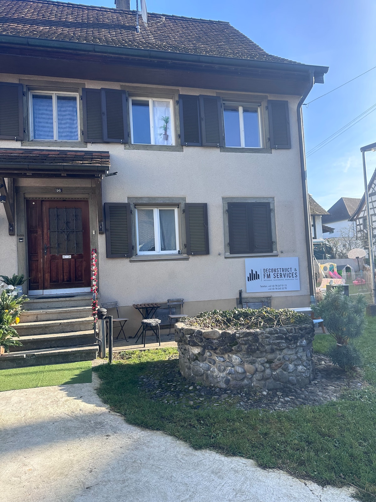 A two-story building is presented with an inviting entrance. The facade features wooden shutters and a wooden door. A small stone garden bed is located in front, surrounded by a well-maintained lawn. A sign for services is displayed prominently near the entrance.