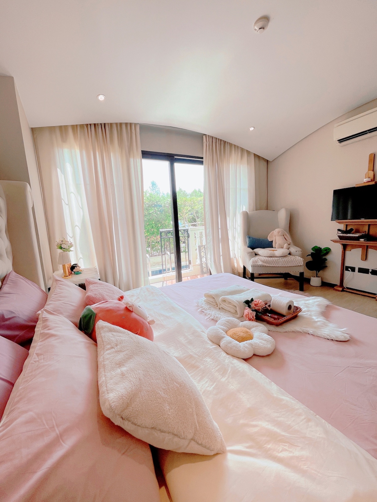 A bright bedroom features a spacious bed adorned with soft pink bedding and plush pillows. Natural light floods the room through large glass doors, which open to a balcony. A comfortable armchair sits nearby, accompanied by a small plant beside it.