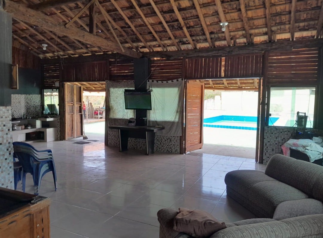 A spacious living area features rustic wooden beams and open doors, allowing natural light to fill the room. A cozy seating arrangement is visible, along with a wall-mounted television. The adjacent area opens to a refreshing pool, enhancing the indoor-outdoor living experience.