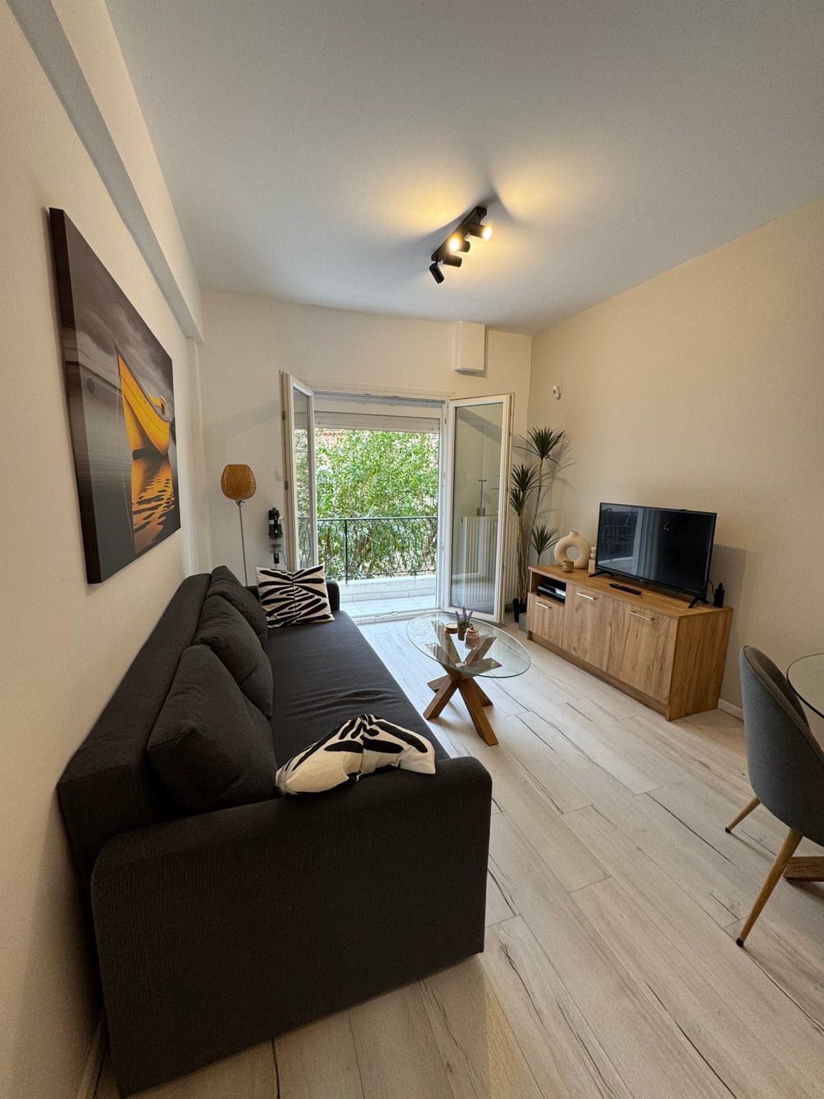 A cozy living room area features a dark gray sofa with decorative cushions. A wooden coffee table is positioned in front, complementing the wooden TV stand. Glass doors open to a small balcony with greenery visible outside, and light fixtures enhance the space.