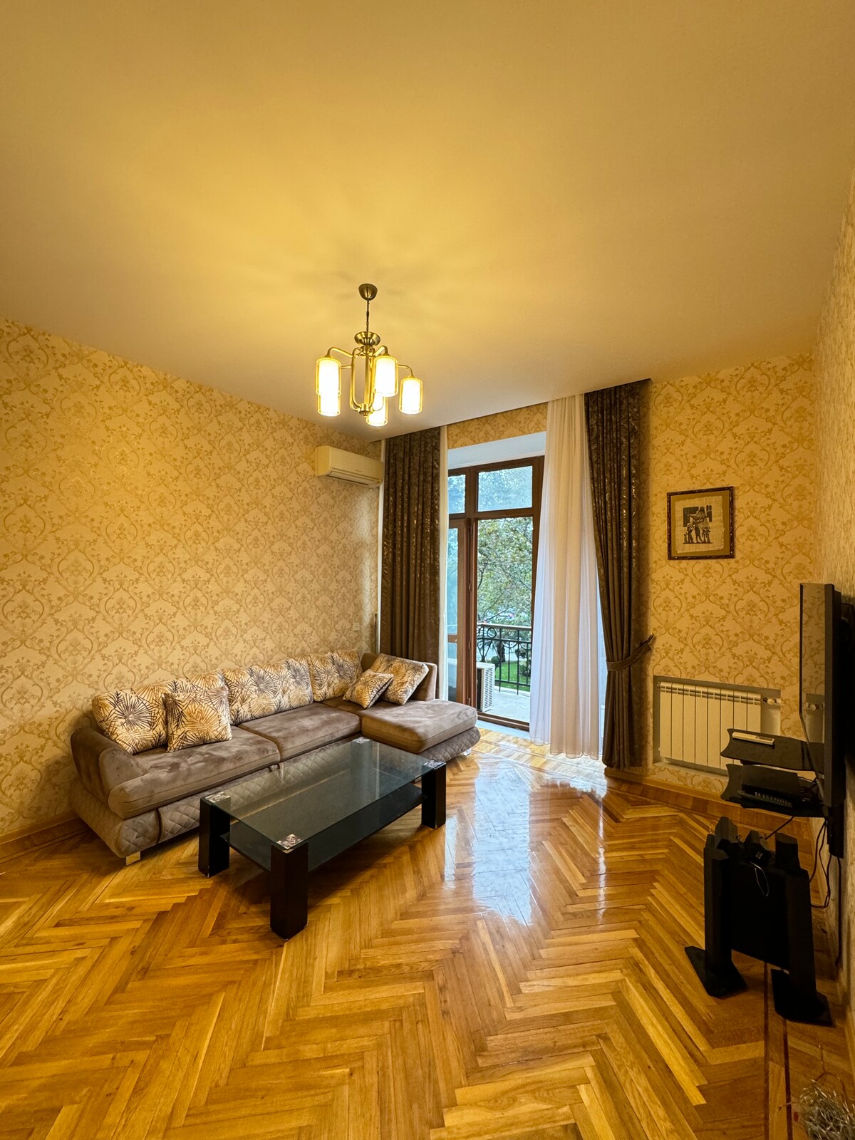 A cozy living area is furnished with a sectional sofa adorned with patterned cushions. A glass coffee table rests on a herringbone wooden floor, while a flat-screen television is positioned nearby. Natural light enters through double doors leading to a balcony, framed by long curtains.