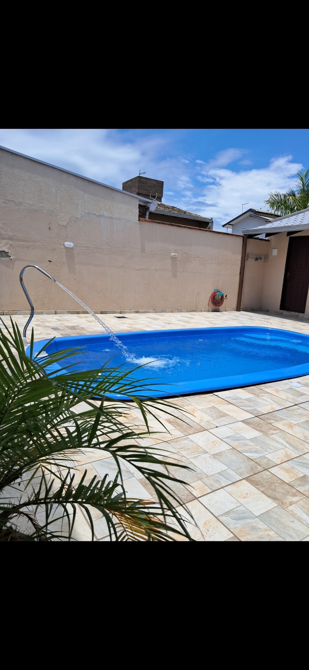 A serene outdoor area features a blue swimming pool surrounded by light-colored stone tiles. A lush green plant is positioned in the foreground. The space is illuminated by natural light, with clear skies and a few clouds visible overhead.