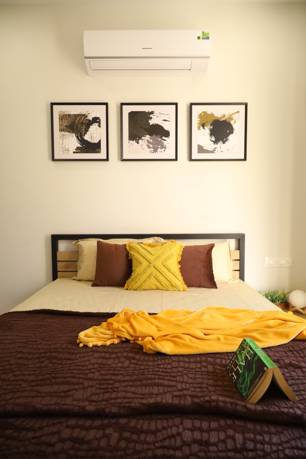 A cozy sleeping area features a spacious bed adorned with a patterned brown coverlet. Bright yellow throw blankets and a decorative pillow add color. Above the bed, three framed artworks are displayed, and an air conditioning unit is mounted on the wall.
