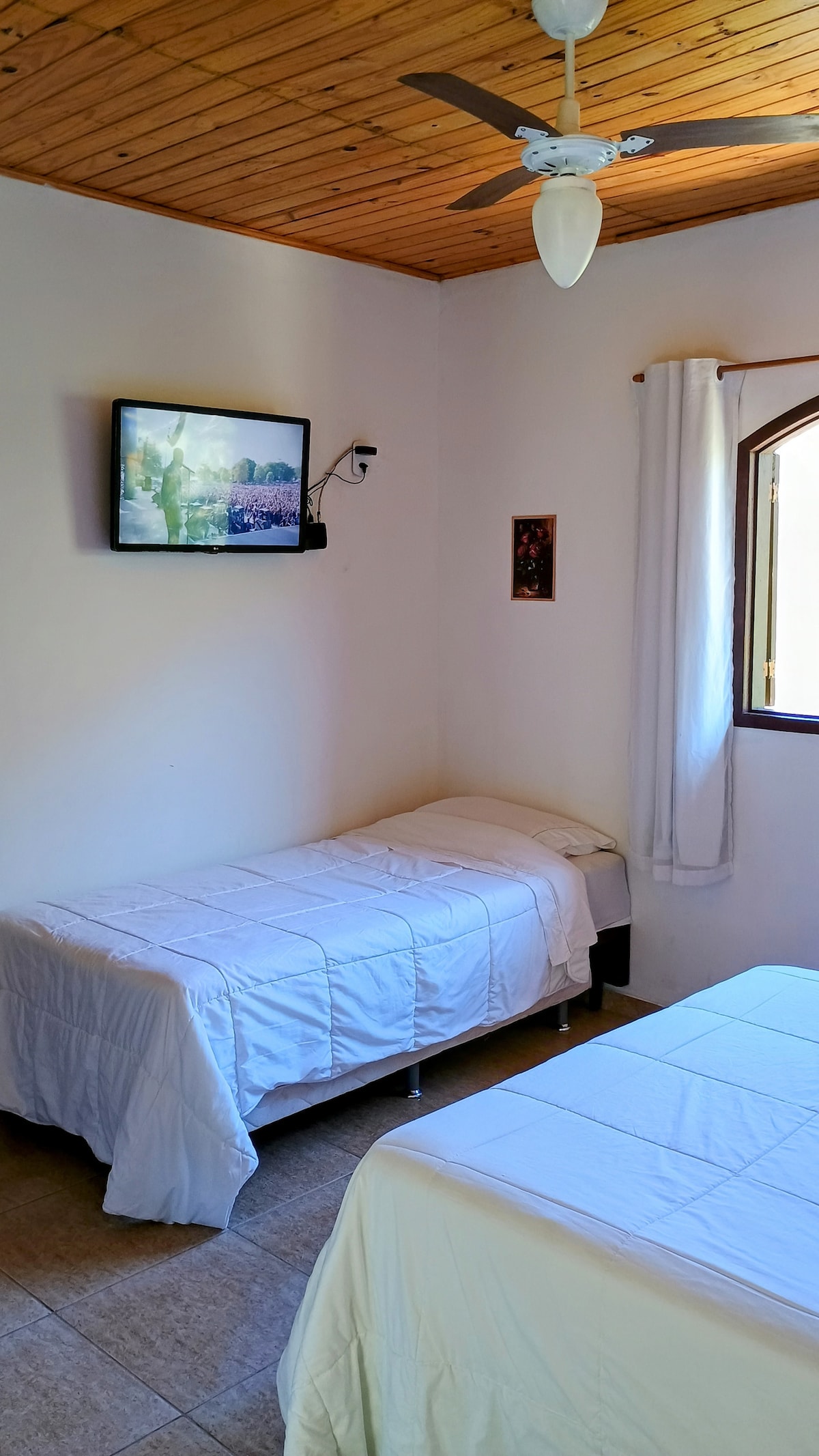 A cozy room features two beds with white bed linens positioned side by side. A mounted television is visible on the wall opposite the beds. Natural light enters through a window adorned with a light curtain, while wooden ceiling accents complement the inviting space.