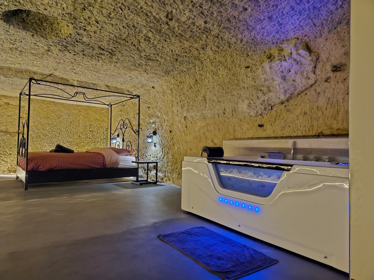 An expansive love room features a four-poster bed, complemented by soft bedding. A modern whirlpool bathtub, accentuated with blue lighting, sits nearby against natural stone walls, highlighting the room's unique ambiance. The floor is smooth, enhancing the minimalist aesthetic of the space.