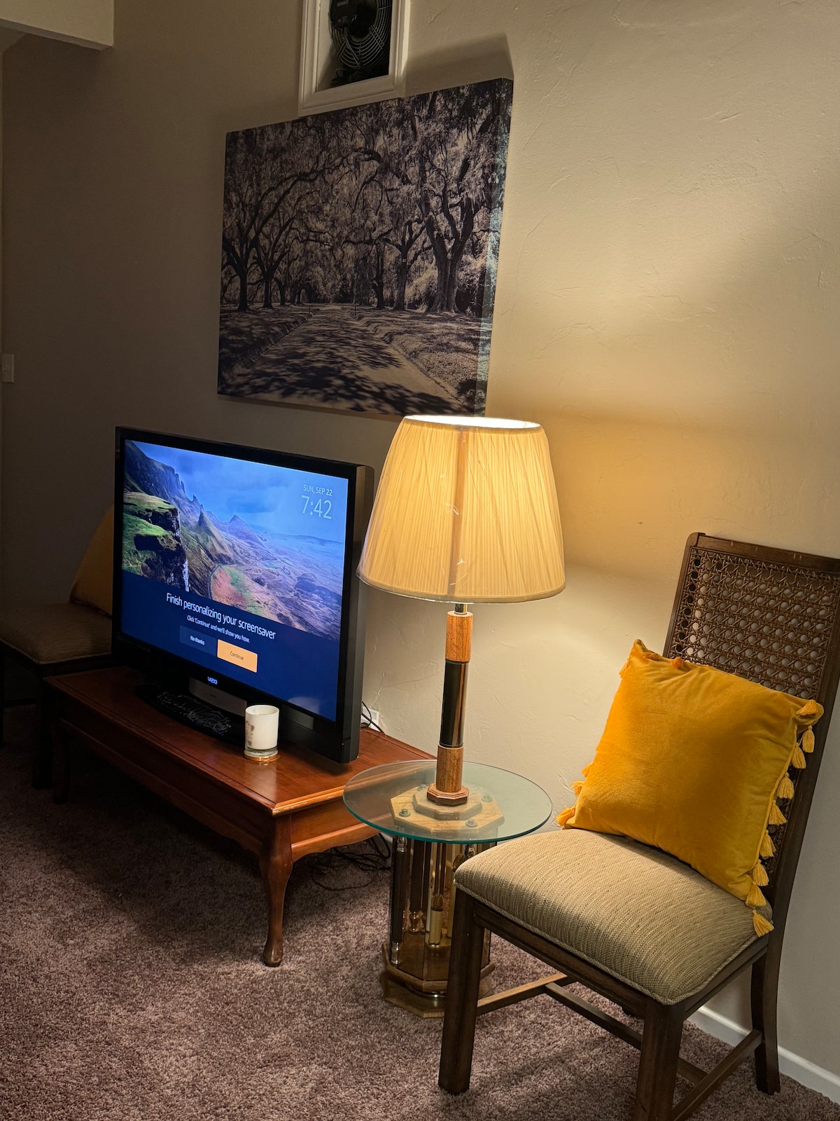 A cozy corner of the living room is highlighted by a television displaying a serene landscape. Nearby, a lamp emits soft light beside a chair with a decorative yellow pillow. A wooden table holds a candle, contributing to the warm atmosphere.