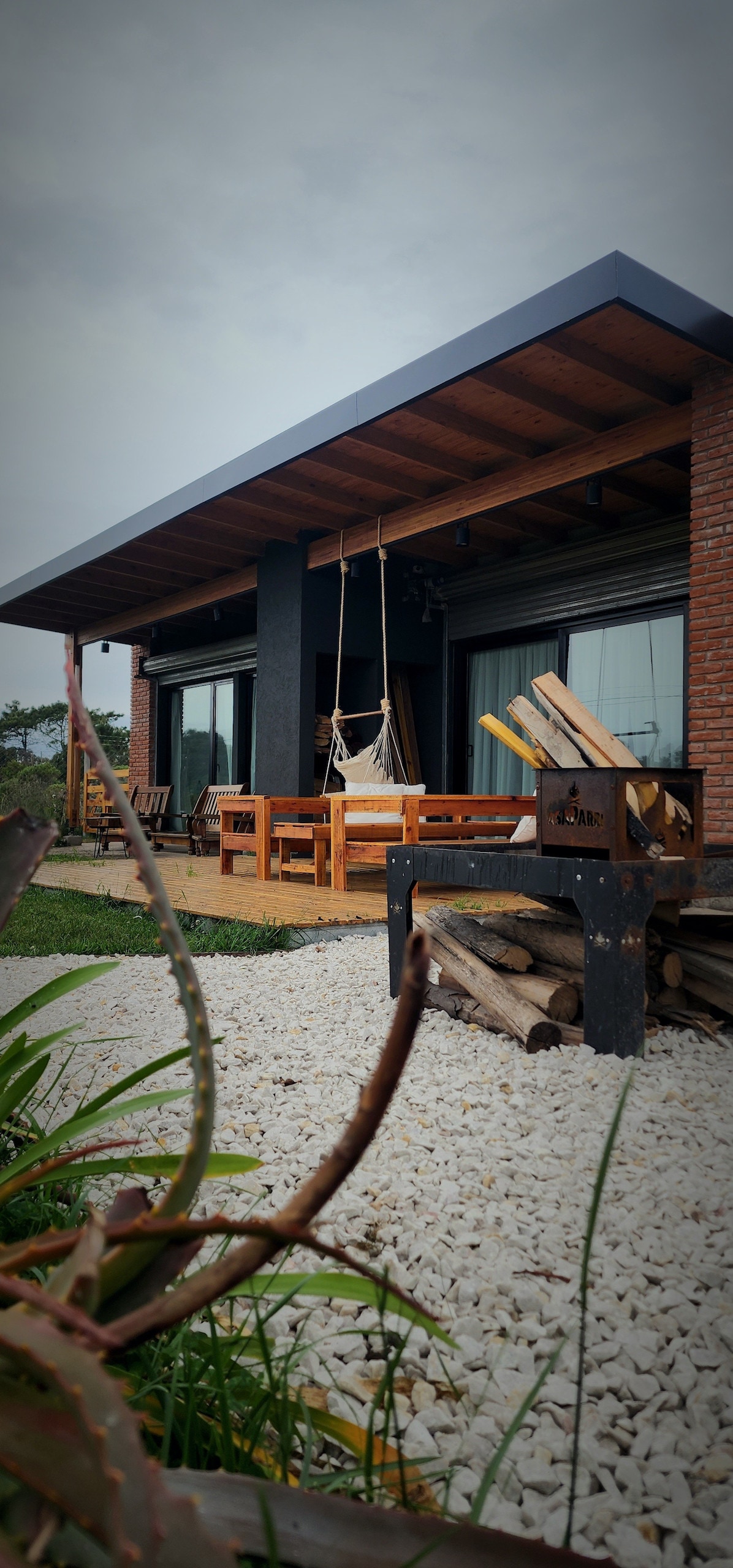 The outdoor area features a covered patio with comfortable seating and a wooden table, inviting relaxation. An outdoor grill is positioned nearby, alongside a stack of firewood. Surrounding greenery and gravel pathways enhance the natural ambiance of the space.