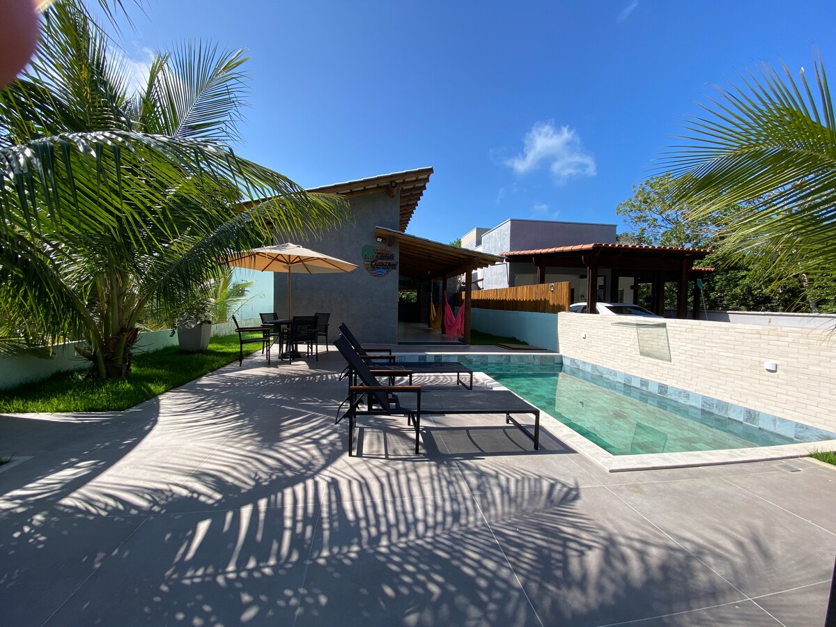 A serene outdoor area features a turquoise pool surrounded by lush greenery and palm trees. Sun loungers are positioned near the water, and a shaded dining area with an umbrella provides a space for relaxation. The backdrop includes charming architectural elements against a clear blue sky.