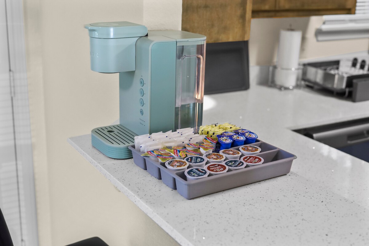 A modern coffee maker in a soft blue hue is positioned on a light countertop. An array of coffee and tea pods is neatly organized in a tray beside the machine, offering various flavors. Natural light illuminates the kitchen space softly.