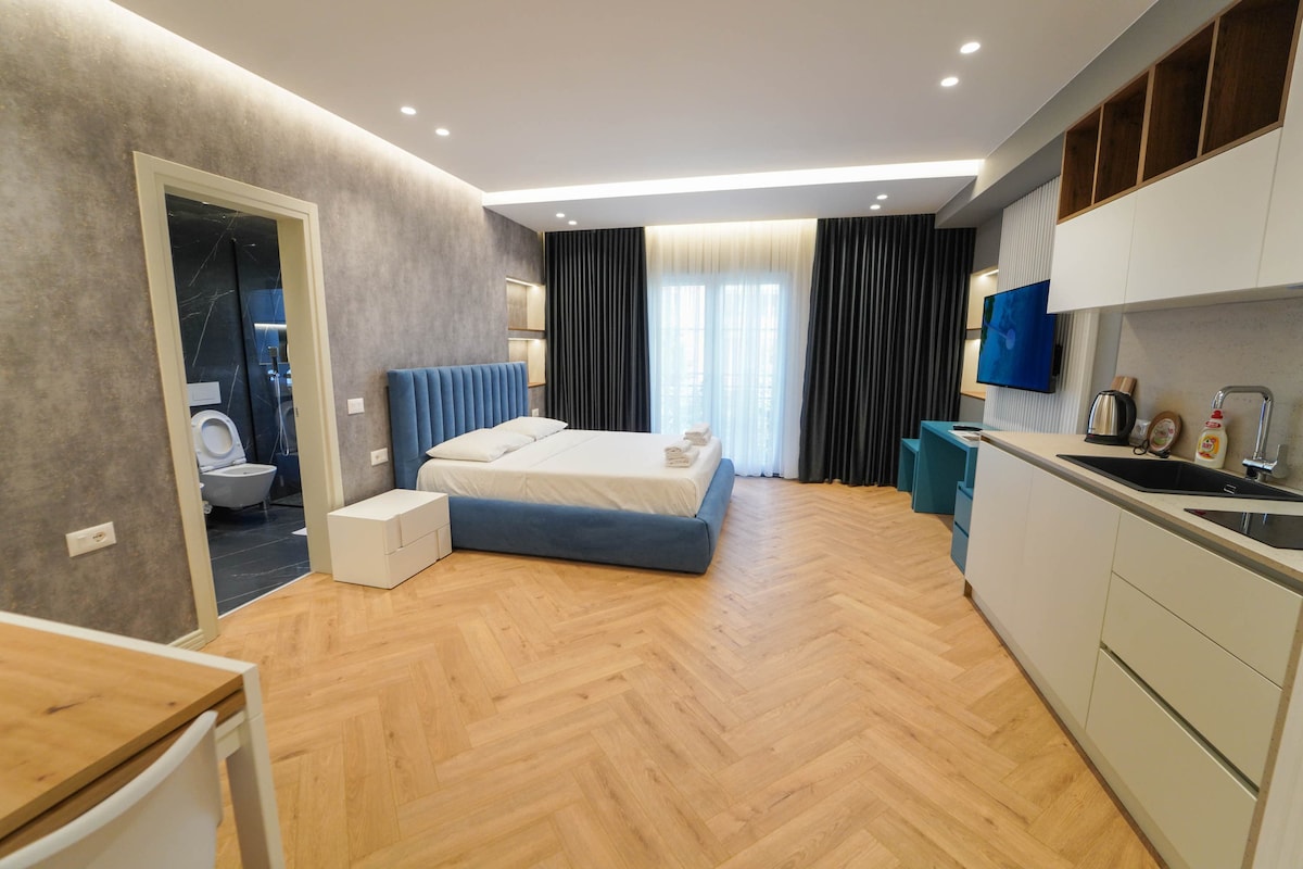 A modern studio apartment features a spacious bed with crisp linens against a soft grey wall. Natural light enters through large windows, highlighting the wood flooring. An open kitchenette with minimalist cabinetry and contemporary appliances is visible, while a bathroom door is partially open in the background.