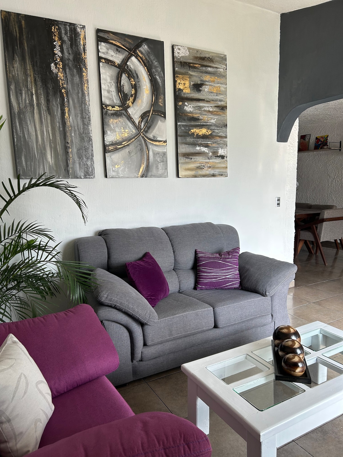 A cozy living area is seen featuring two sofas, one in gray and another in vibrant purple. Above, three abstract art pieces in gold and black are displayed on the wall. A coffee table with a decorative centerpiece adds to the inviting atmosphere.