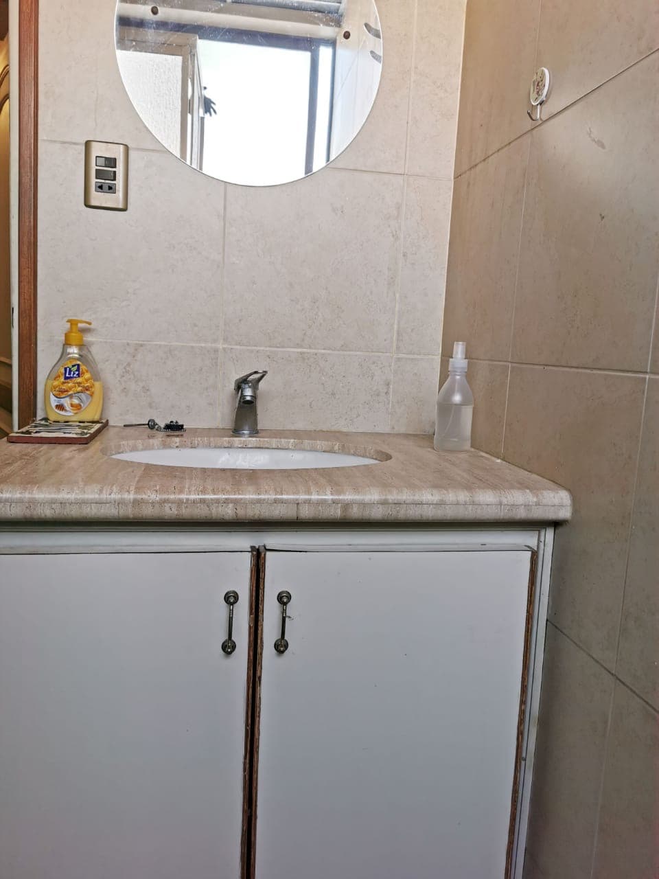 A bathroom countertop is visible, featuring a circular mirror above. The sink is positioned on the left side, complemented by a faucet. A cleaning product and a spray bottle are placed on the surface. The cabinet beneath provides storage space.