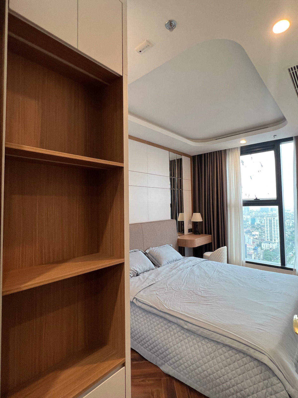 A well-lit bedroom features a spacious bed with neutral bedding. A wooden shelf is visible to the left, providing storage space. Large windows allow natural light to fill the room, while city views can be seen in the background. Soft curtains frame the windows.