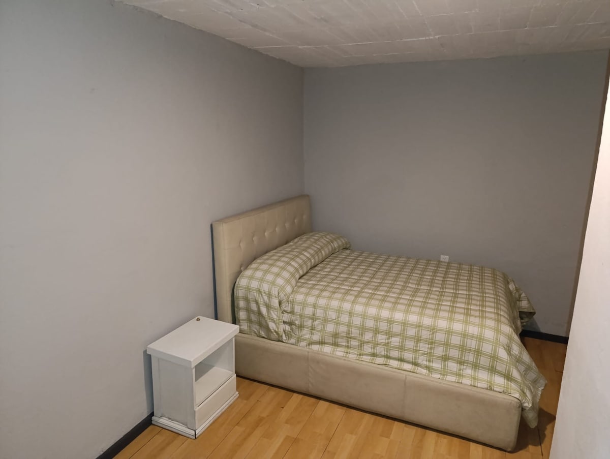 A cozy sleeping area is presented with a light-colored bed featuring a padded headboard, covered with a green and white checkered blanket. A small nightstand with a clean design is positioned beside the bed. The walls are painted in a soft gray, and the flooring is light wood.