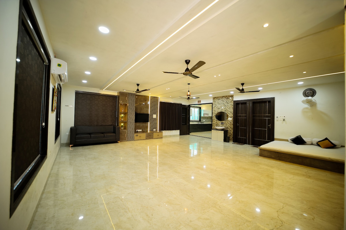 A spacious living area features polished marble flooring with multiple lighting options. Ceiling fans are present for comfort. A dark sofa sits against one wall, while a cozy seating area is positioned near a window, offering a view of the surrounding space. Elegant wall textures add depth.
