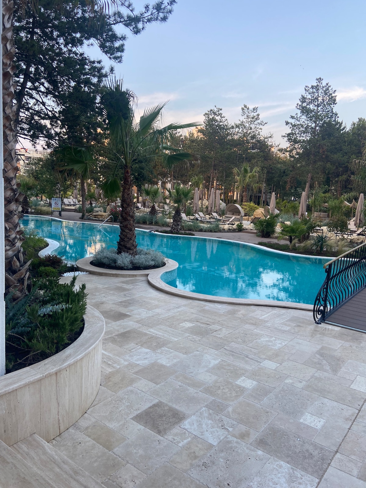 A large swimming pool is framed by lush palm trees and landscaped greenery. The pool's clear blue water reflects the surroundings, while a pathway of square stone tiles leads to the water's edge, inviting relaxation and leisure.