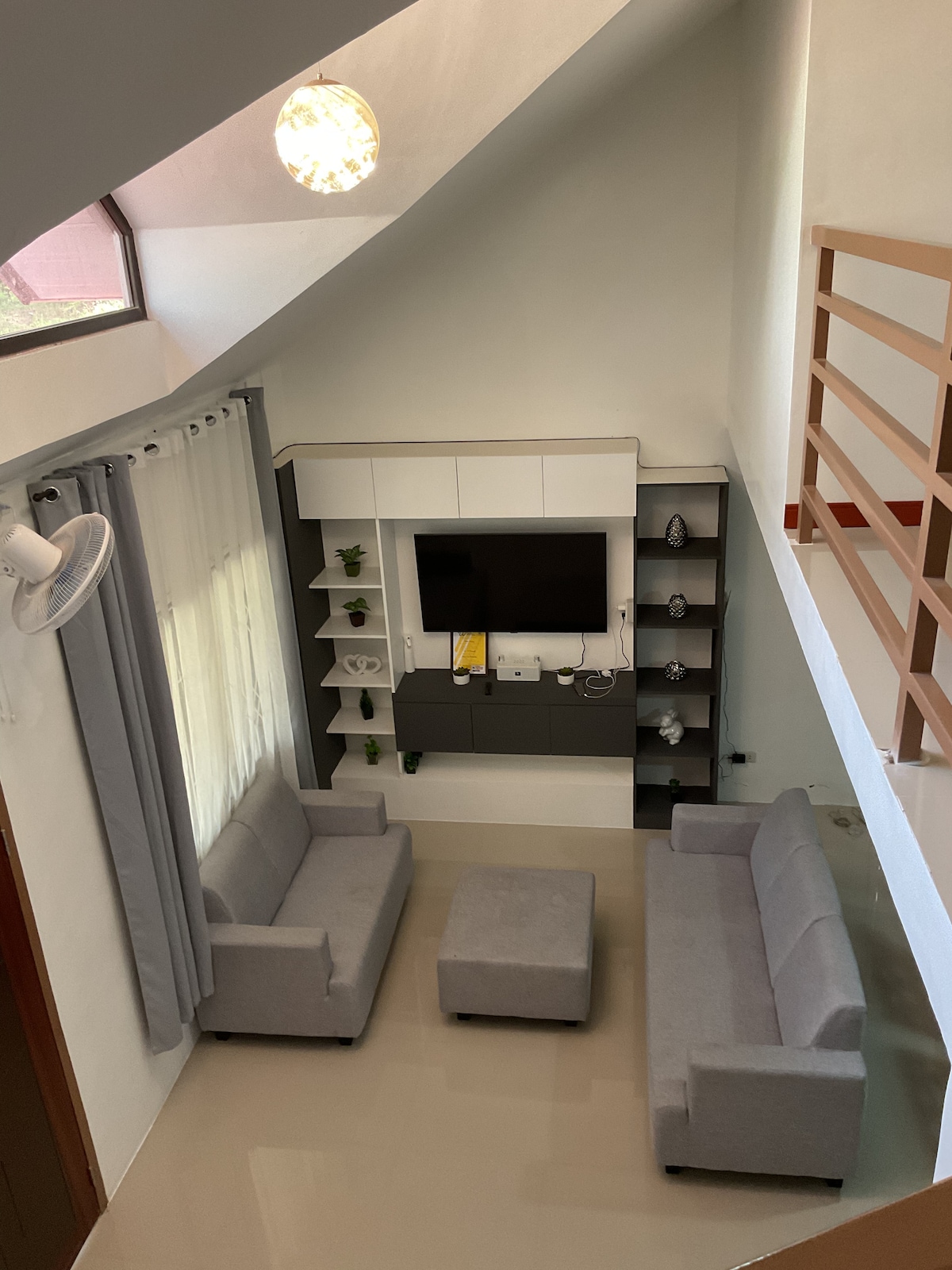 A sunlit living area is viewed from above, featuring two light gray sofas arranged around a small, matching coffee table. A modern entertainment unit with decorative plants is positioned against a white wall. Natural light filters through large windows, and soft gray curtains frame the space.