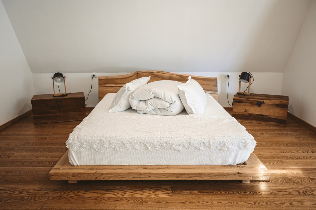 A spacious bed with white linens and several plush pillows is positioned on a wooden platform. Wooden nightstands are located on either side, complemented by lantern-style lamps. The smooth hardwood floor adds warmth to the serene room.