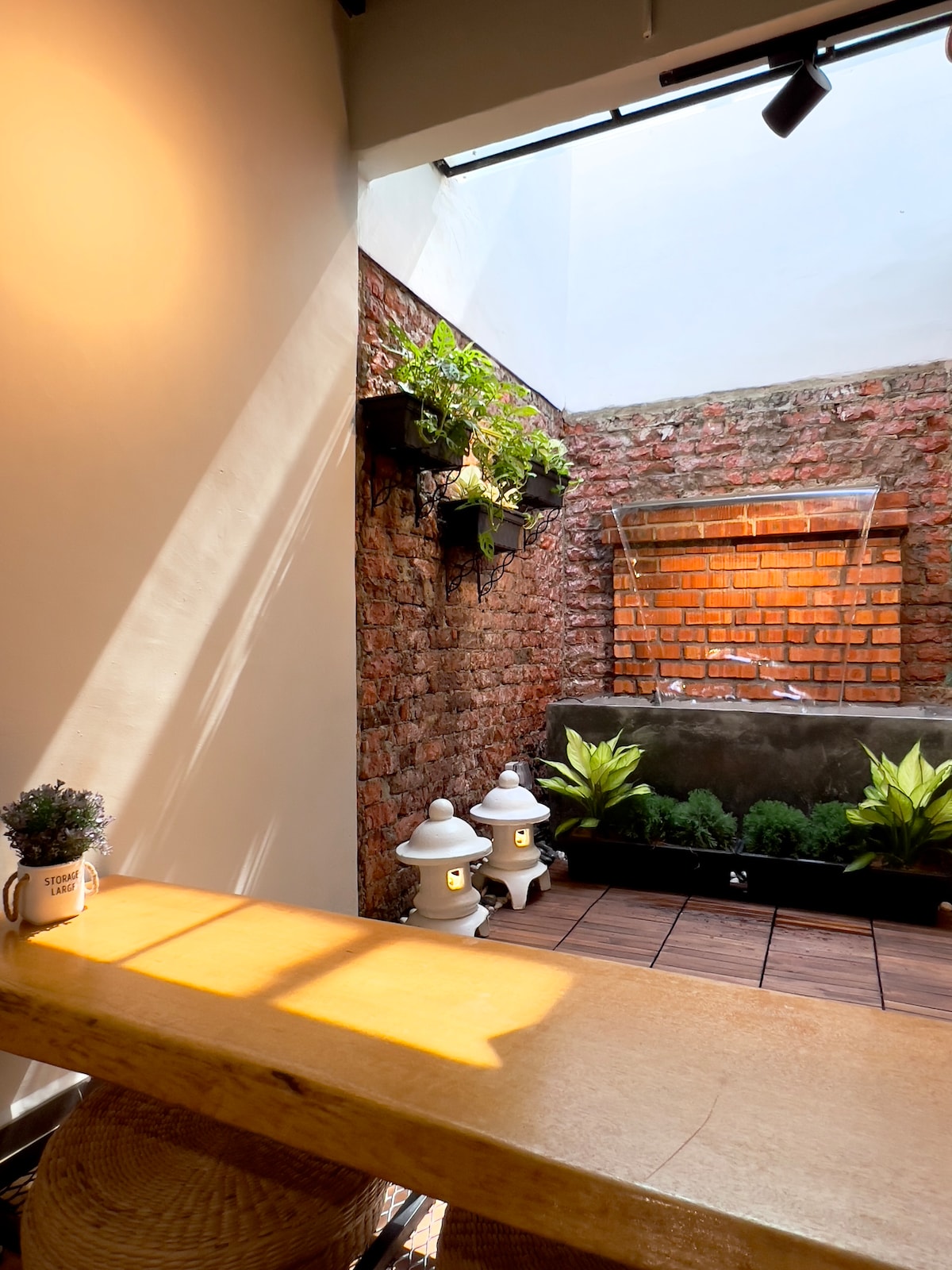 An interior courtyard features exposed brick walls adorned with potted greenery. A gentle water feature creates a serene atmosphere, while two lanterns provide soft illumination. Natural light filters through the space, enhancing the inviting ambiance.