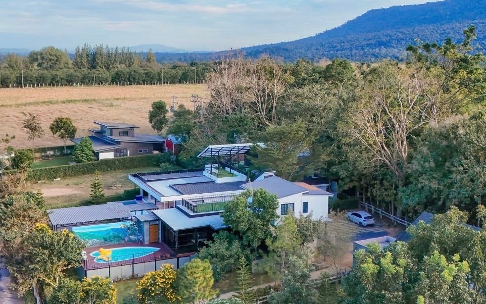 An aerial view showcases a spacious two-story private villa surrounded by lush greenery and mountains. The property features a sparkling blue pool and a rooftop area for stargazing, complementing its serene outdoor environment.