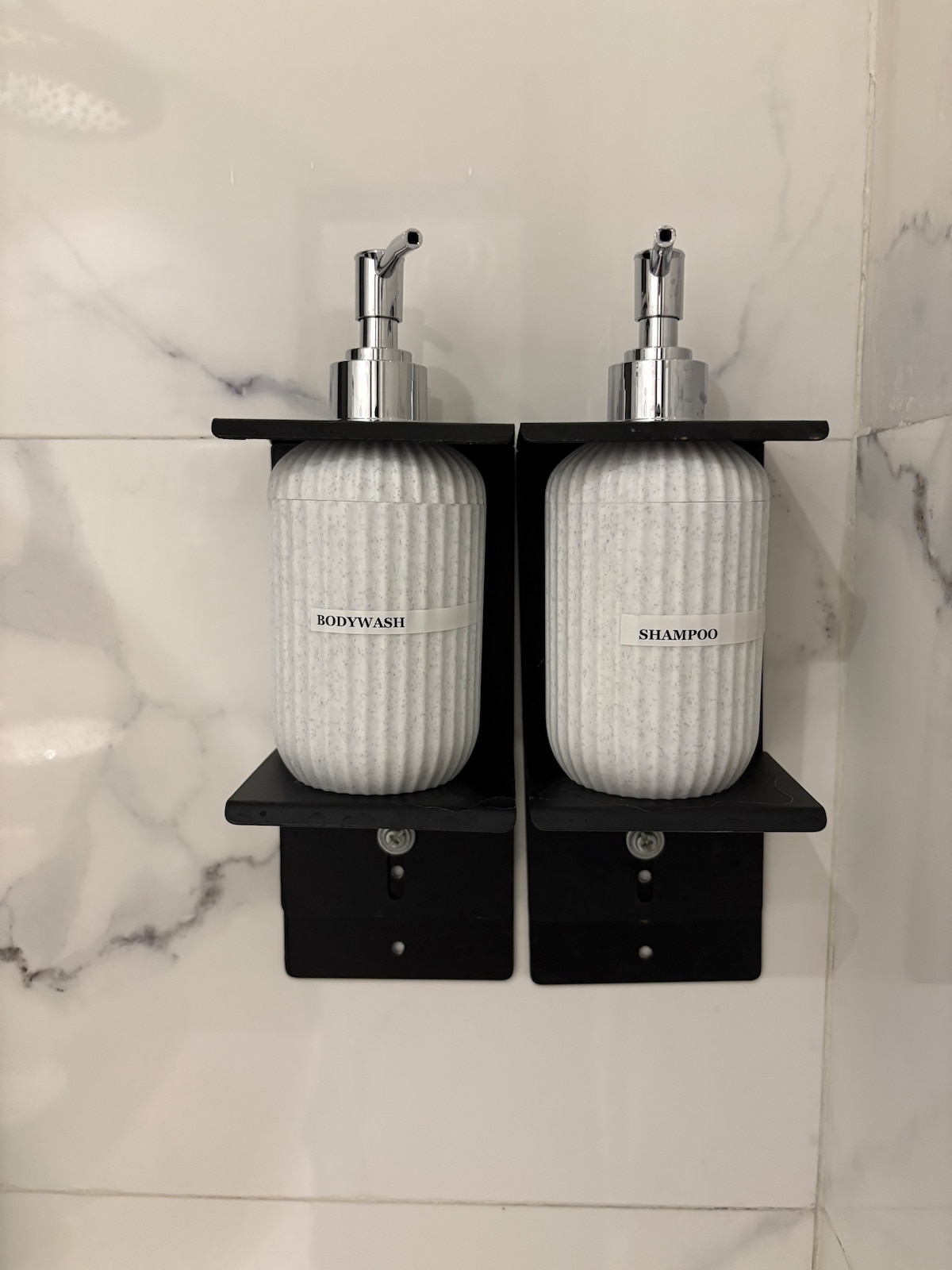 A shower area features two wall-mounted dispensers, one labeled for body wash and the other for shampoo. The dispensers have a textured white design, with a sleek silver pump. The background consists of white marble tiles, creating a clean and modern appearance.