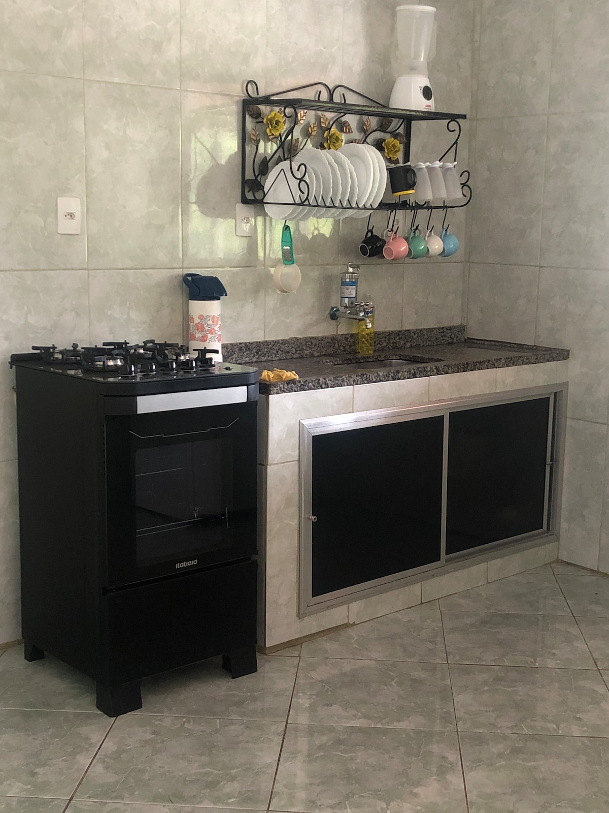 A compact kitchen features a black gas stove and a countertop with a silver fronted cabinet. Shelves above display neatly arranged dishes and colorful mugs. Natural light brightens the space, enhancing the light-colored tile walls.