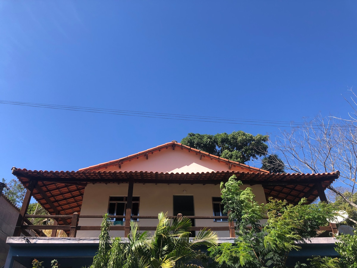 The exterior of the house is framed by vibrant green trees and tropical plants. A terracotta-tiled roof and wooden balcony railings contribute to its rustic charm. A clear blue sky provides a bright backdrop, creating a relaxed and inviting atmosphere for potential guests.