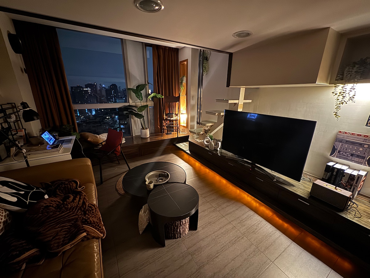 A cozy living area is illuminated with warm lighting, featuring a large TV mounted on a sleek unit. A brown sofa sits beside two round coffee tables made of wood, while large windows reveal a nighttime city skyline. Potted plants add a touch of nature.