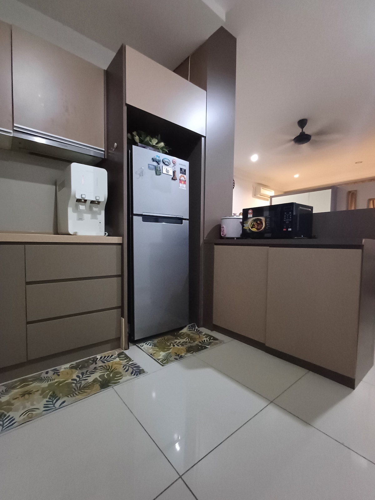 The kitchen area features modern cabinetry and sleek surfaces. A water dispenser is positioned next to the refrigerator, which is situated against a neutral wall. A microwave and an induction cooker are visible on the countertop, complemented by decorative mats on the tiled floor.