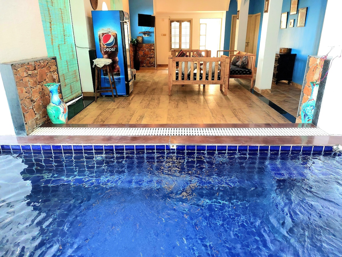 A view of a serene indoor pool reflects the blue tones of the adjacent living area. Light wooden flooring can be seen, leading to a seating area with a wooden sofa and decorative elements visible in the background.