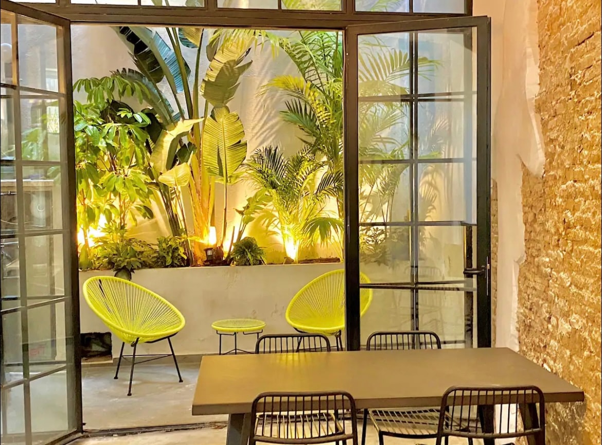 A private patio is visible through large glass doors, featuring vibrant greenery and soft lighting. Two bright yellow chairs and a small table create a cozy seating area. A simple dining table with black chairs occupies the foreground, enhancing the space's inviting ambiance.