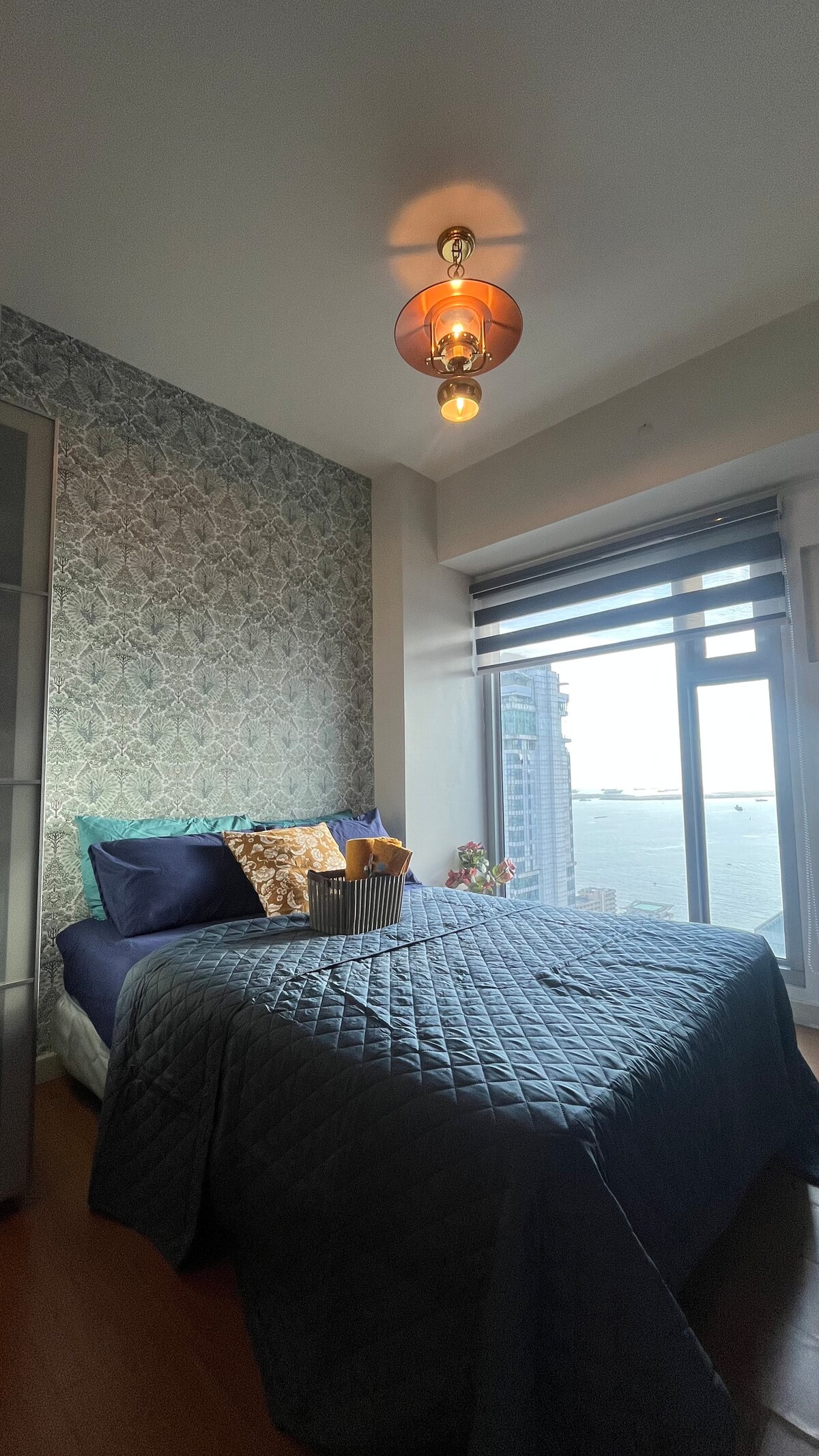 A cozy sleeping area features a spacious bed dressed in a dark blue quilt, complemented by decorative pillows. Large windows reveal expansive views of the sea, while a patterned accent wall adds a touch of detail. Soft light emanates from a stylish ceiling fixture.