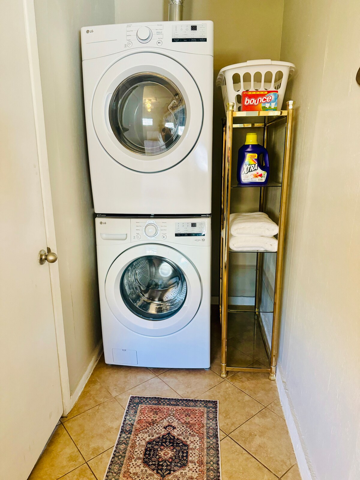 A laundry area is presented, featuring a stacked washer and dryer in white. A gold shelving unit holds rolled towels, laundry supplies, and a basket. The floor is tiled, and a small, decorative rug is placed in front of the appliances.